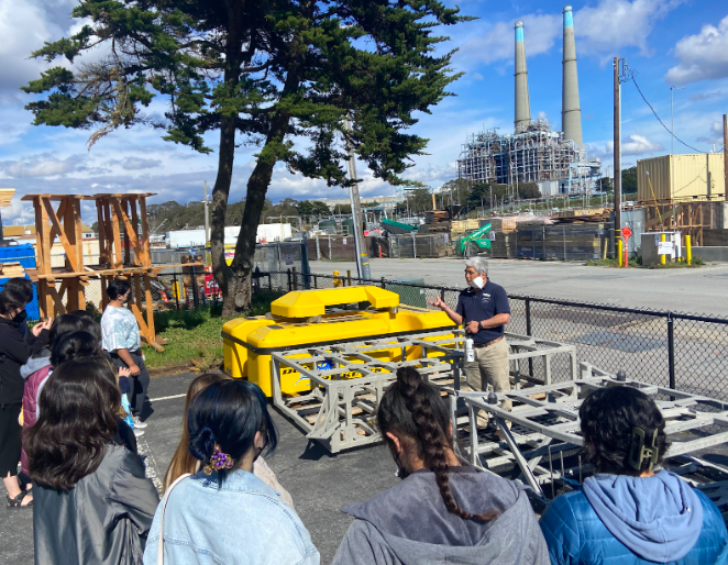 Students and instructor standing in lots with factory in background.